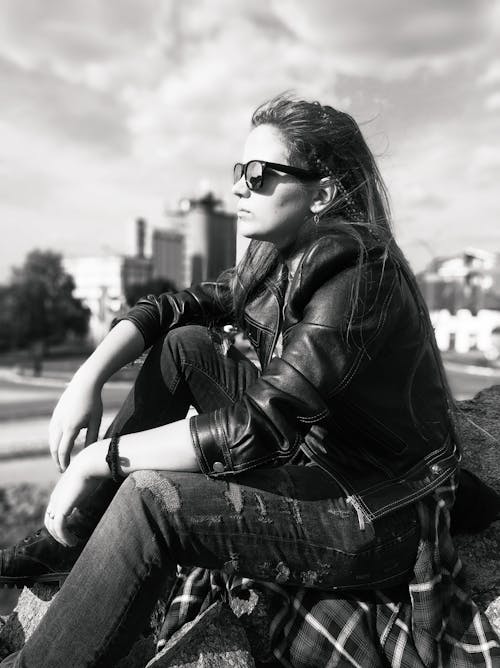 Black and white side view of contemplative female in trendy leather jacket and sunglasses sitting on staircase in city while looking away