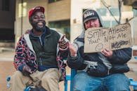 Cheerful black man in cap sitting near plump tattooed male with SMILE AT LEAST YOUR NOT ME inscription on cardboard while showing with fingers at each other on street