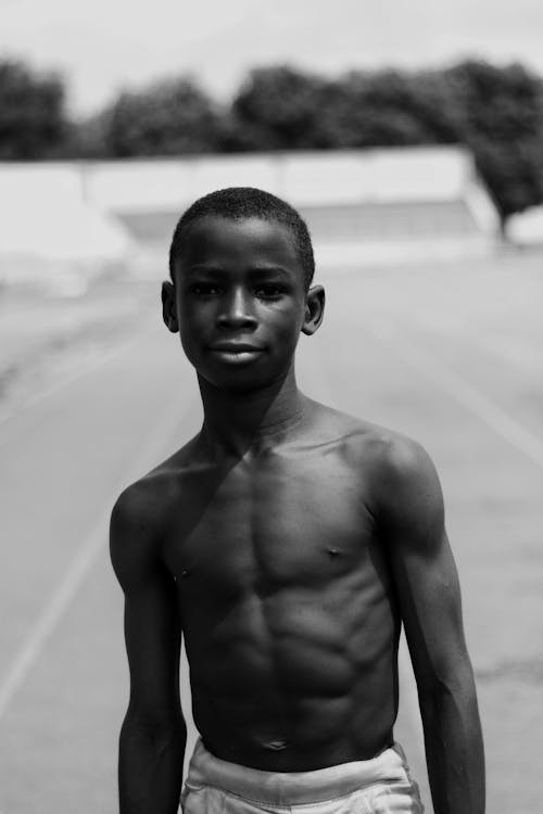 Black and white of muscular African American teen with naked torso looking at camera while standing on asphalt roadway