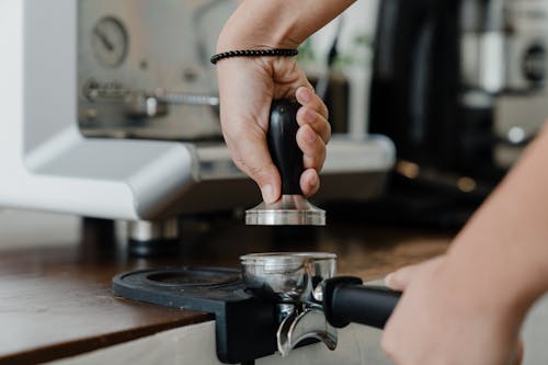 Unrecognizable female bartender pressuring ground coffee in portafilter with tamper for preparing tasty aromatic espresso during work in coffee house