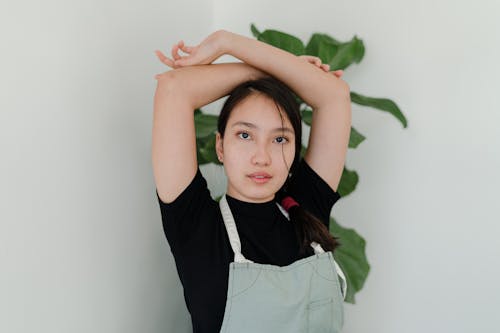 Positive young Asian female worker wearing apron standing with raised and crossed arms against wall and potted plant in modern shop during work time while looking at camera