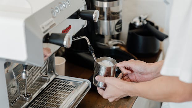 Unrecognizable female bartender preparing milk foam for cappuccino or latte while working with coffee machine at wooden counter in modern coffee house