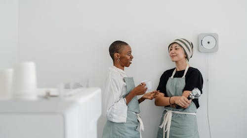 Couple of multiracial female baristas wearing apron standing near wall in modern coffee shop with minimal interior while drinking coffee and communicating and looking at each other