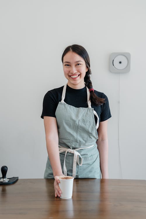 Free Positive young Asian woman wearing apron giving paper cup with coffee to go while standing at wooden counter during work in modern coffee house and looking at camera with smile Stock Photo