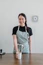 Positive young Asian woman wearing apron giving paper cup with coffee to go while standing at wooden counter during work in modern coffee house and looking at camera with smile