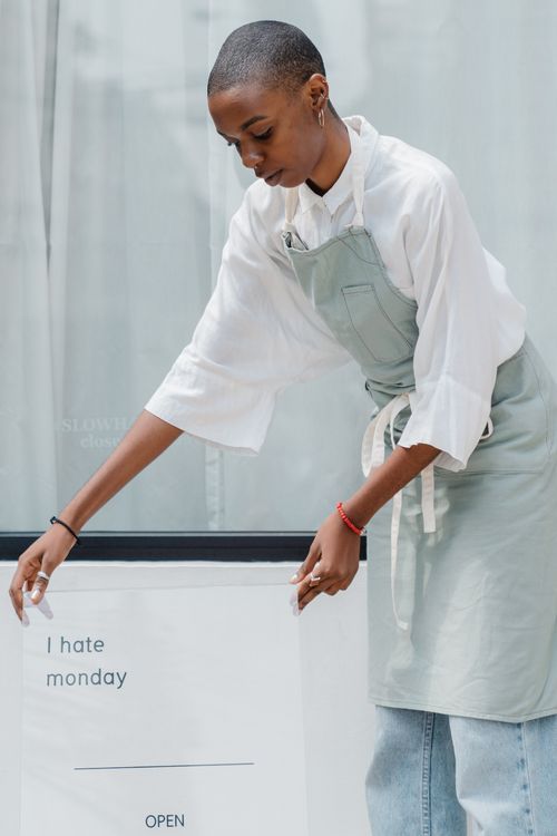 Free Young African American woman with short hair wearing apron setting transparent information banner with I HATE MONDAY inscription and working hours near window Stock Photo