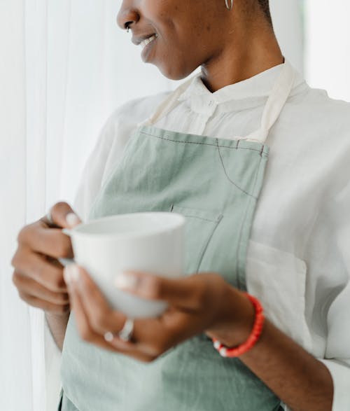 Smiling black female barista in apron standing with ceramic cup of coffee in hands during worktime in coffee house