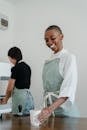 Happy young black woman with short hair wearing apron serving paper cup with coffee to go while standing at wooden counter and looking down with smile