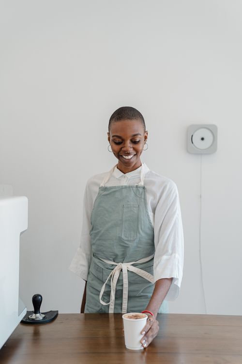 Free Happy young black woman with short hair wearing apron giving paper cup with coffee to go while standing at wooden counter during work in modern coffee house and looking down with smile Stock Photo