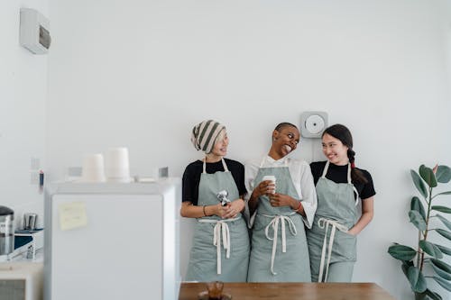 Multiracial female baristas wearing apron standing together near wall in modern coffee shop with minimal interior during workday and looking at each other with smile