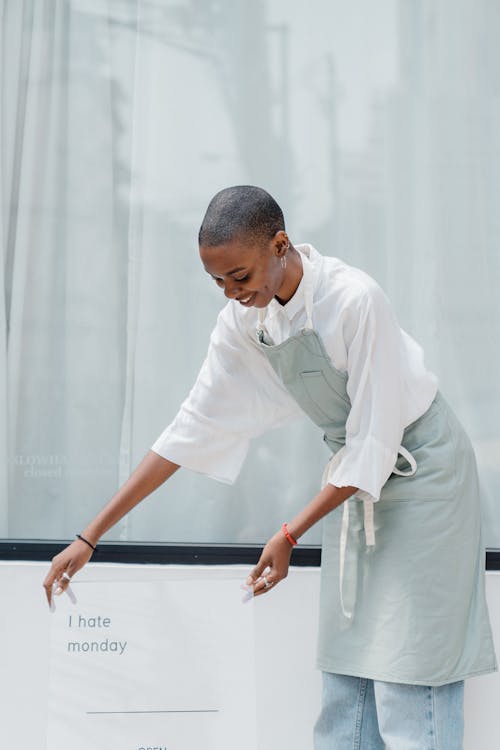 Free Young African American woman with short hair wearing apron putting information banner with I HATE MONDAY shop name and working hours near window of store or cafe Stock Photo
