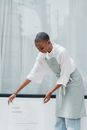 Young African American woman with short hair wearing apron putting information banner with I HATE MONDAY shop name and working hours near window of store or cafe