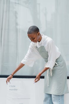 Young African American woman with short hair wearing apron putting information banner with I HATE MONDAY shop name and working hours near window of store or cafe