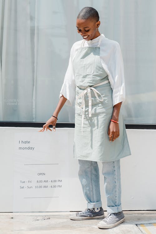 Full body smiling young African American woman with short hair in apron standing near transparent banner with working hours on sunny street