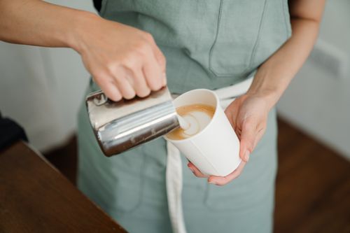 Free Unrecognizable female in apron pouring hot milk into paper cup while preparing aromatic latte to go in modern coffee house Stock Photo