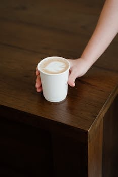 From above of unrecognizable female bartender putting paper cup with delicious latte to go on wooden counter while working in modern coffee house