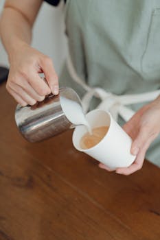 Barista skillfully pouring steamed milk into a coffee cup, creating a foamy latte art in a café setting.