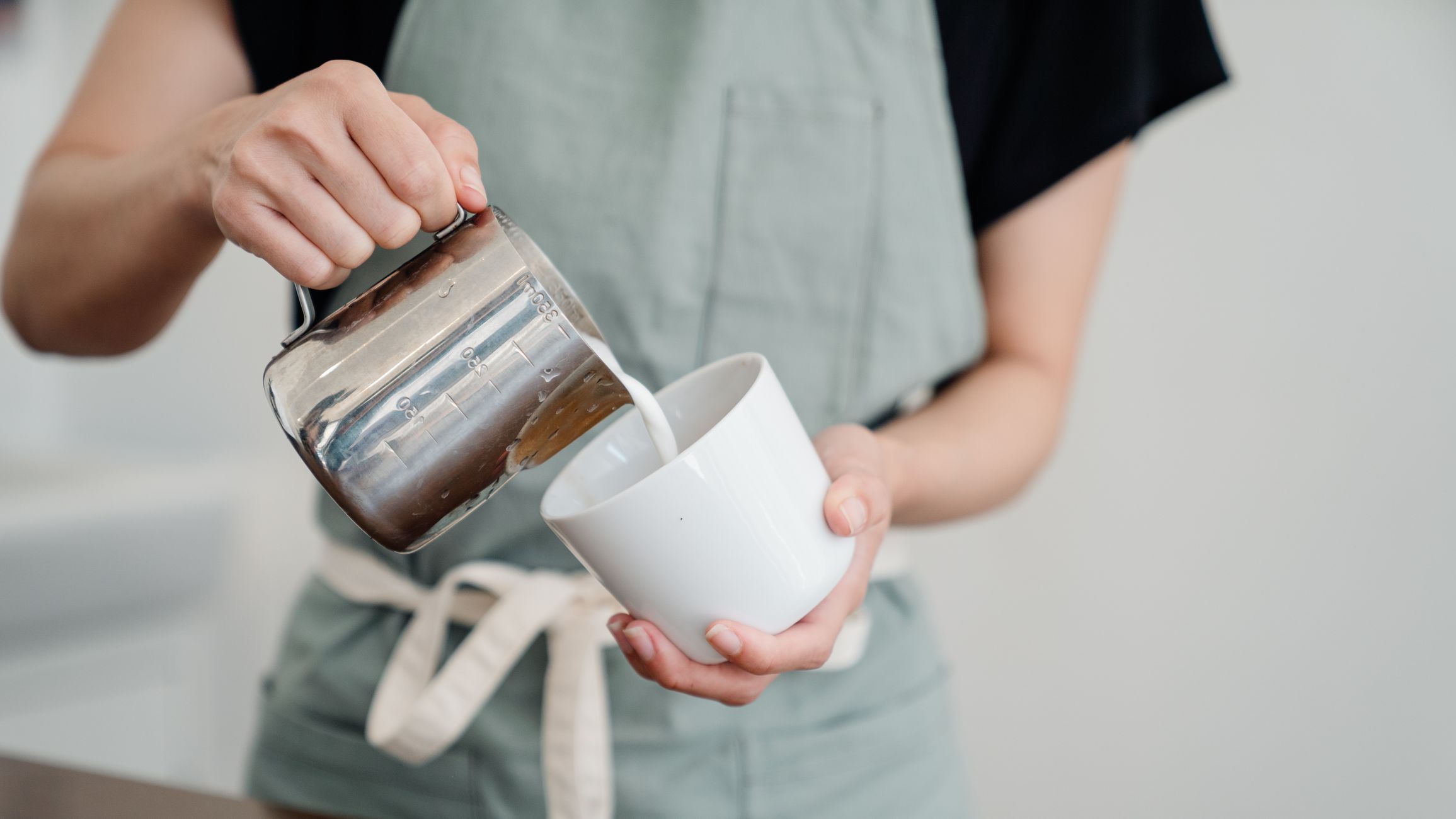 Unrecognizable barista in apron pouring milk into ceramic cup while preparing aromatic coffee in modern cafe