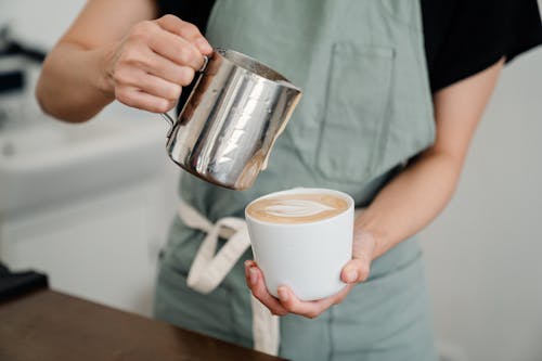 Crop bartender in apron preparing aromatic cappuccino in cup while pouring foam of milk into ceramic cup in coffeehouse