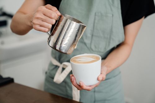 Free Crop bartender in apron preparing aromatic cappuccino in cup while pouring foam of milk into ceramic cup in coffeehouse Stock Photo