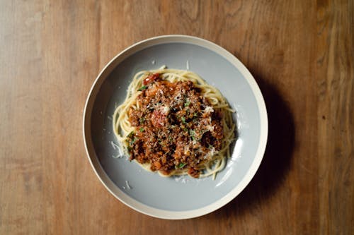Top view of round plate with delicious Italian pasta Bolognaise garnished with grated parmesan cheese placed on wooden table