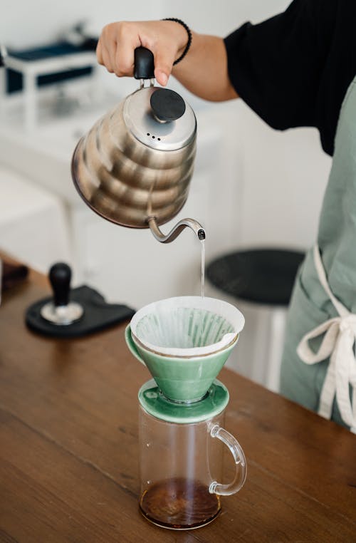Crop anonymous barista in apron using ceramic funnel dripper and metal kettle and disposable filter and glass cup for making pour over coffee