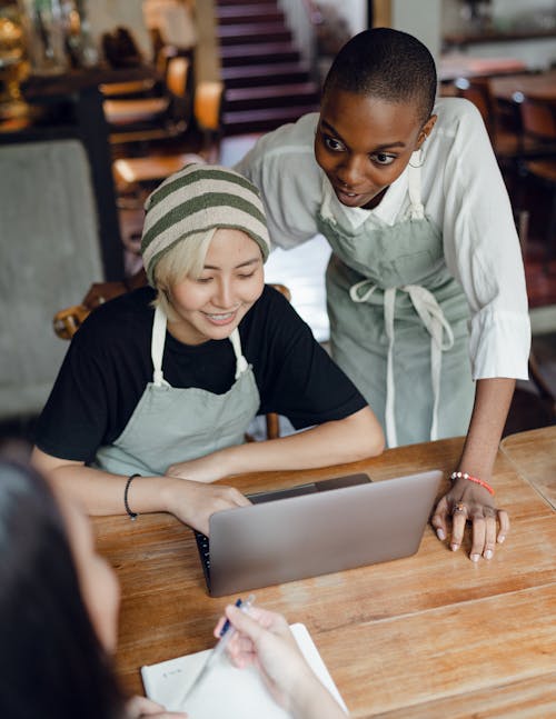 Free From above of optimistic diverse female workers of cafeteria discussing business plan while using laptop and taking notes in piece of paper at table Stock Photo
