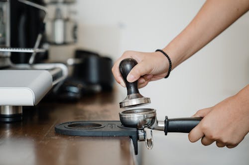 Crop anonymous barista pressing aromatic coffee beans into filter block by using tamper while standing in light kitchen nearby professional brewing machine