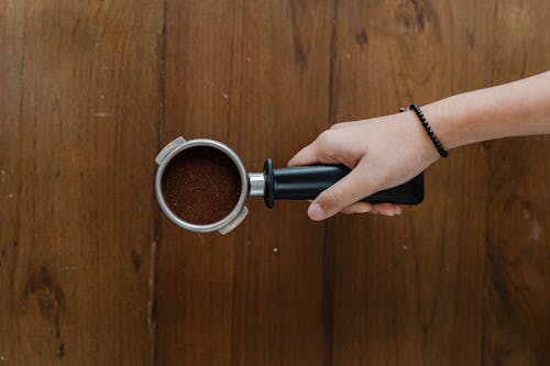 Top view of crop unrecognizable barista hand with metal portafilter full of aromatic ground coffee against wooden shabby table