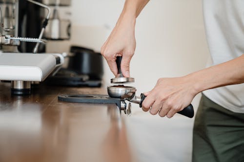 Free Crop anonymous male barista in casual clothes pressing ground coffee into metal portafilter by using tamper while working in contemporary kitchen Stock Photo