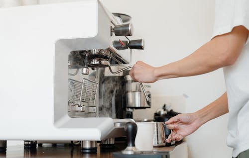 Free Side view crop anonymous barista in white shirt preparing to steam milk in frothing pitcher using professional coffee machine steam wand Stock Photo