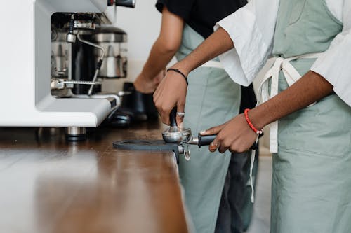 Side view crop anonymous ethnic barista in apron grounding coffee into portafilter and using tamper while working in light modern cafeteria kitchen