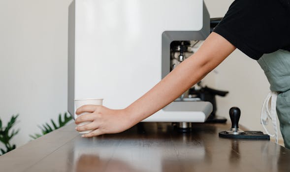 Crop unrecognizable female barista in black shirt and apron standing near modern professional coffee machine and passing white  takeaway paper cup with hot beverage across wooden table in coffee shop