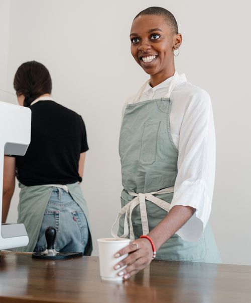 Free Smiling barista holding a cup of coffee behind the counter in a cafe, creating a welcoming atmosphere. Stock Photo