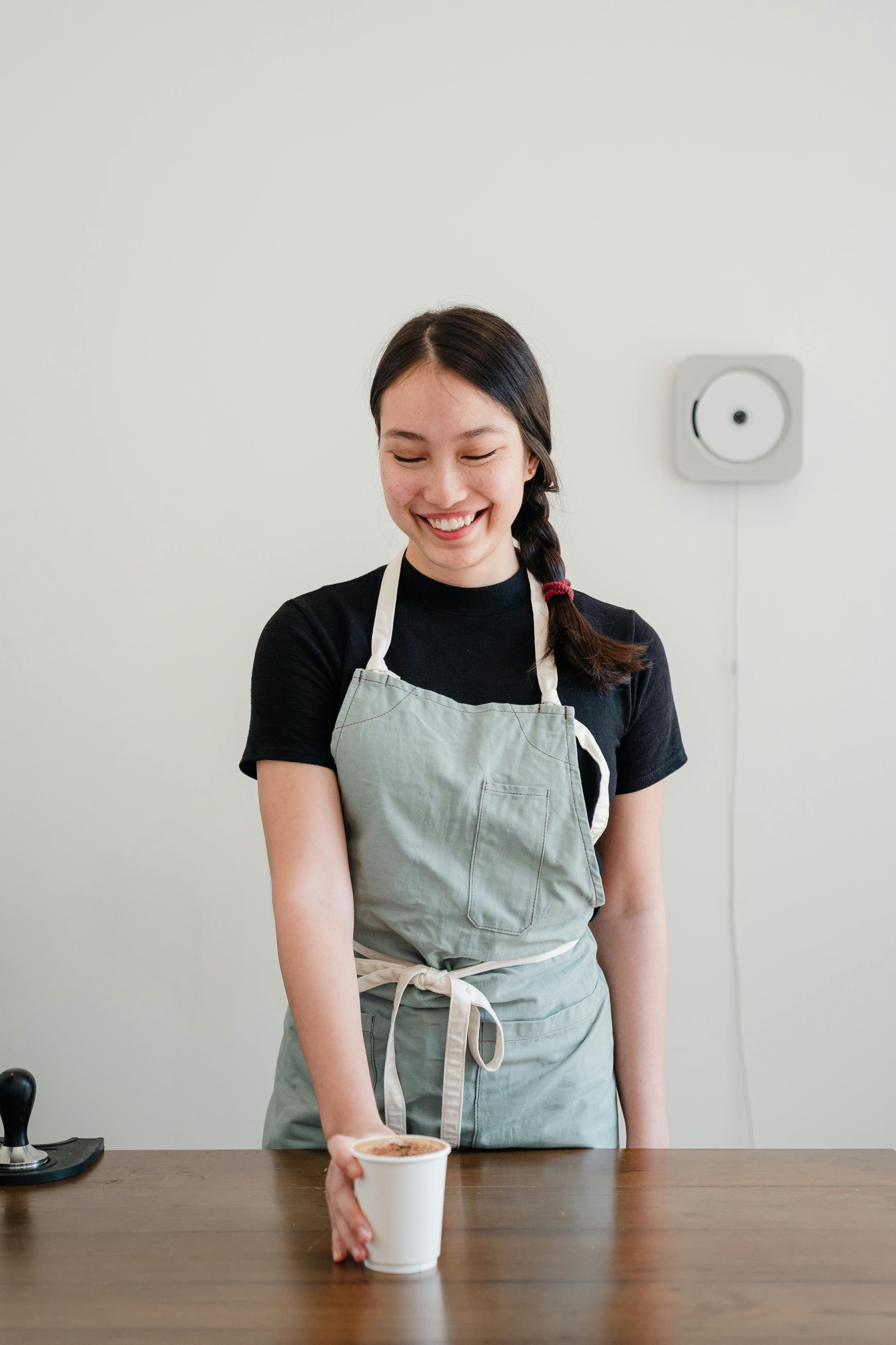 一位开朗的咖啡师系着围裙，在室内端上一杯新鲜咖啡，展现出热情好客