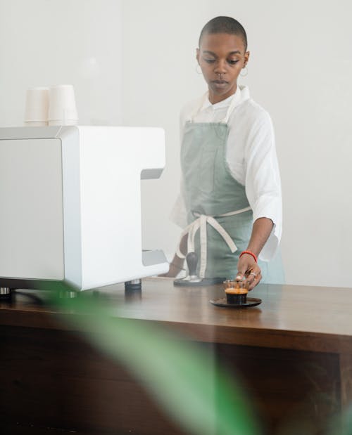 Free Concentrated African American female barista in apron standing nearby professional coffee machine and serving hot aromatic espresso in transparent cup in light cafe Stock Photo