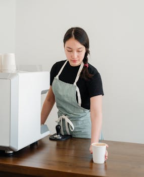 A female barista in an apron serving a cup of coffee in a café with a neutral tone.