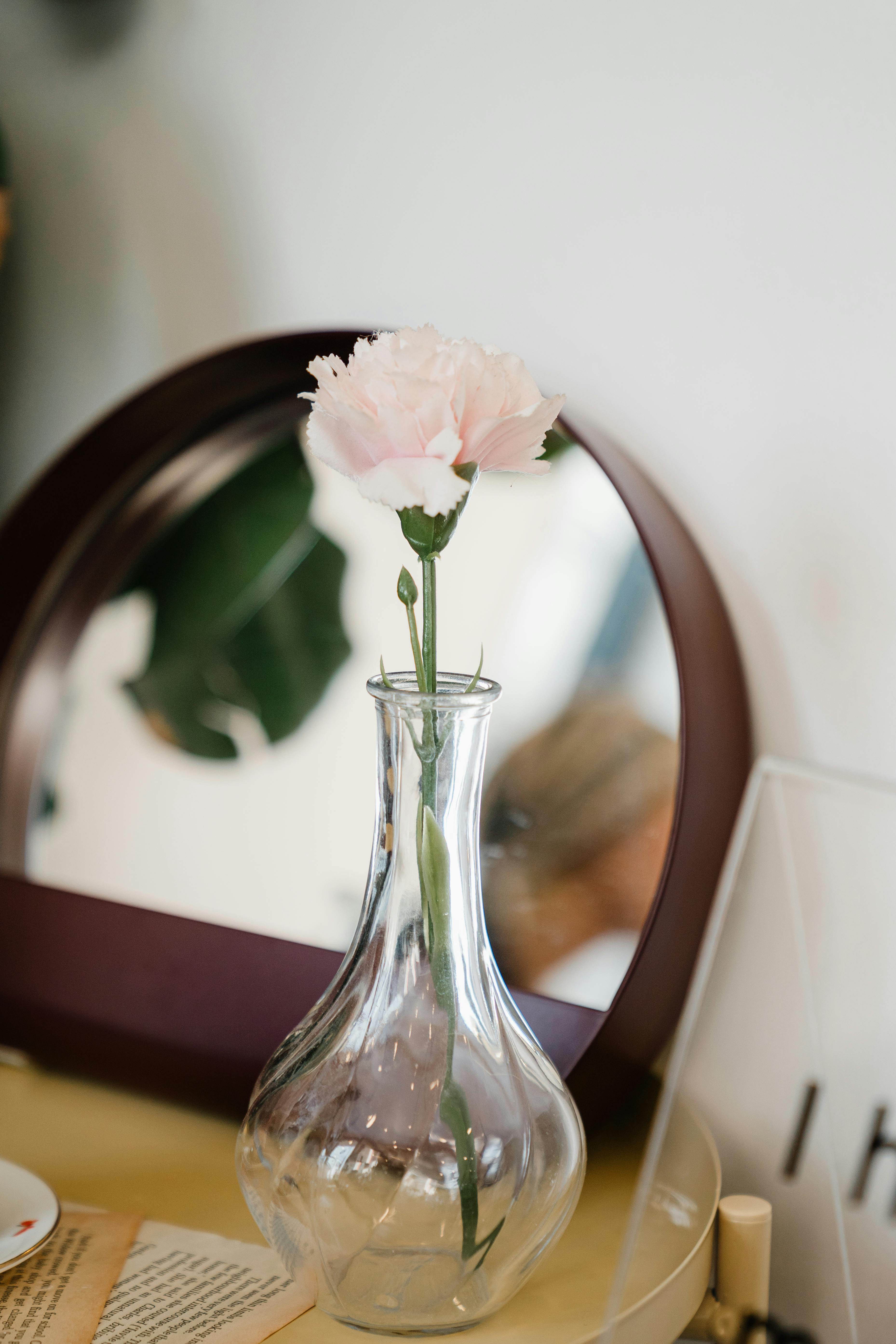 Glass transparent vase with light pink artificial flower placed on wooden table against round mirror in daytime
