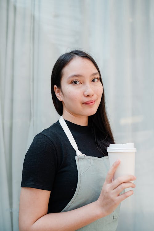 Smiling content Asian female barista in black shirt and apron standing with white takeaway paper cup against blue curtains and looking at camera