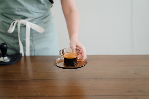 Crop unrecognizable female barista in light green apron passing hot aromatic espresso in stylish transparent cup above wooden table