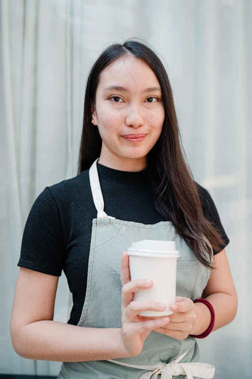 Optimistic smiling Asian waitress with white takeaway paper cup in hands standing against light blue curtains in cozy cafe and looking at camera