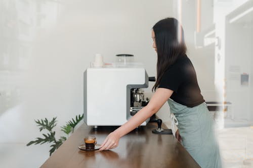 Side view of concentrated female barista in apron standing nearby professional coffee machine and serving freshly brewed aromatic espresso