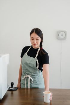 一位围着围裙的年轻少数族裔女咖啡师站在咖啡店柜台前，用纸杯递上一杯卡布奇诺