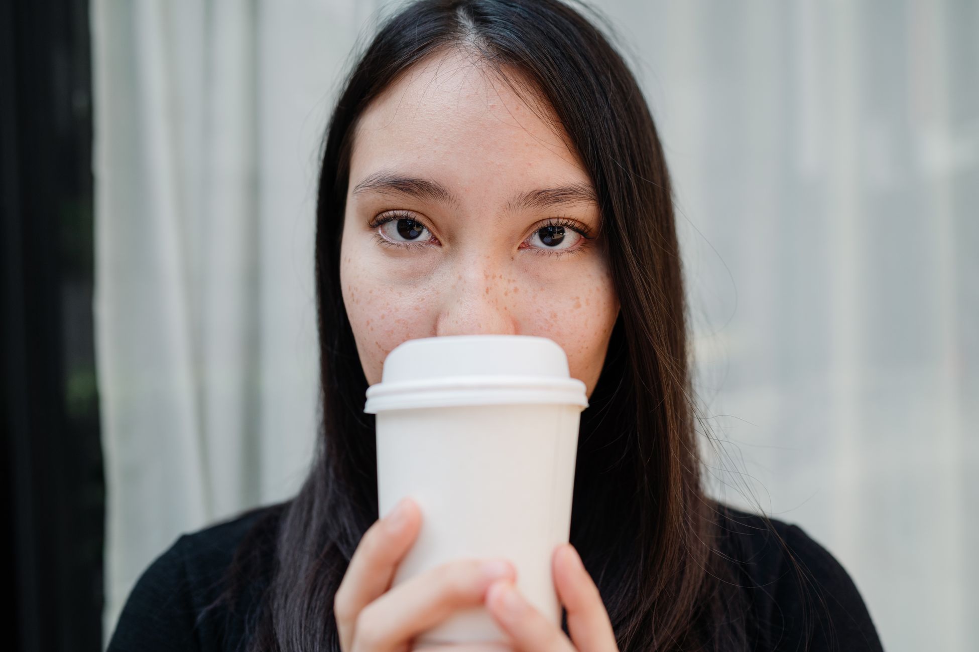 Close-up portrait of a woman sipping coffee from a white disposable cup indoors.