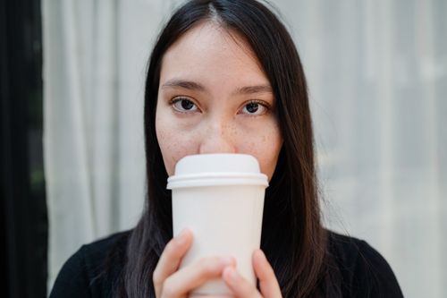 Free Close-up portrait of a woman sipping coffee from a white disposable cup indoors. Stock Photo