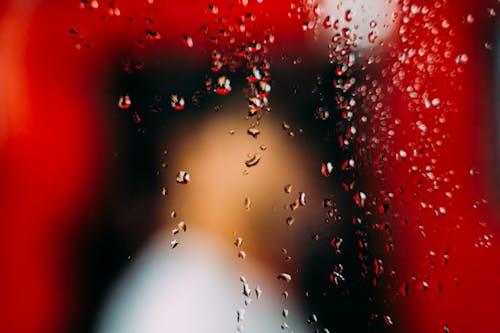 Side blurred view of faceless person standing near bright red construction in form of gate behind window with transparent drips