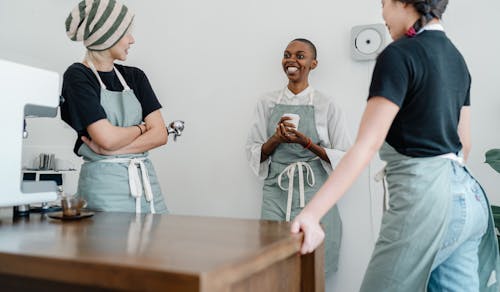 Cheerful young black lady in apron smiling and communicating with colleagues near counter during work in modern coffee shop