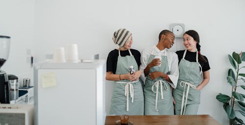 Positive young multiethnic female baristas in aprons talking and smiling while working in modern light cafeteria