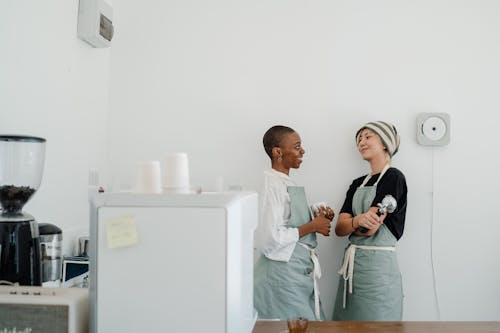 Young smiling multiethnic female baristas in aprons standing leaning on wall and talking during break in cozy coffee shop