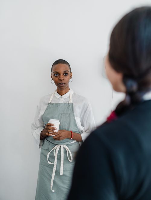 Free A barista in a gray apron holding a coffee cup, engaging in conversation against a white, minimalist background. Stock Photo
